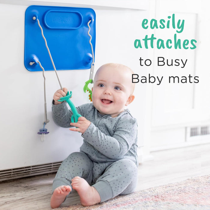 Busy Baby Teething Spoon attached to a busy baby mat suctioned to a refrigerator with text that reads "easily attaches to Busy Baby Mats"