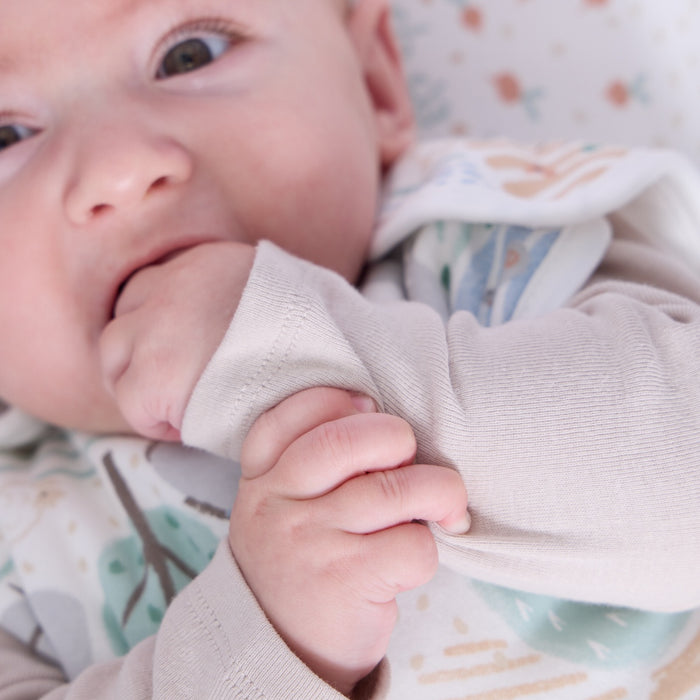 Close-up of baby wearing the Good Life sleep bag, gently chewing sleeve while lying on matching countryside themed sheet
