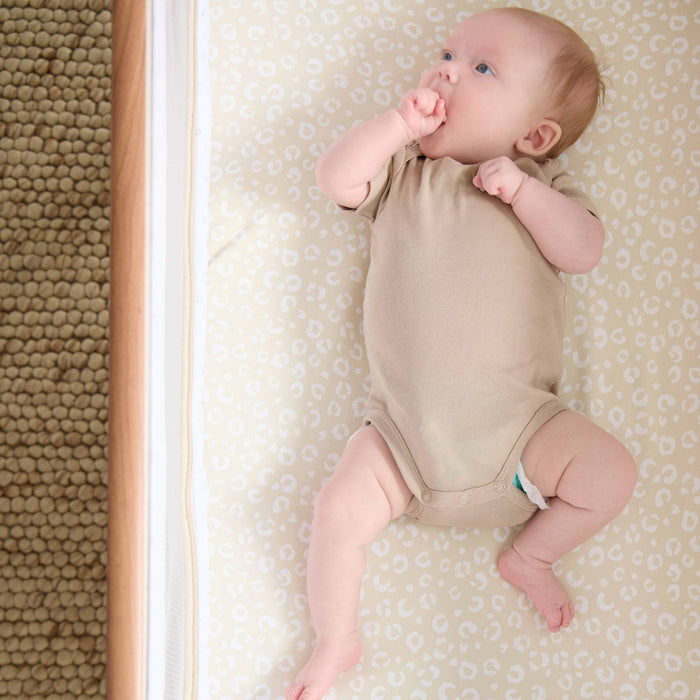 A baby in a beige onesie lying on the CoZee beside crib with the Love Cats fitted sheets