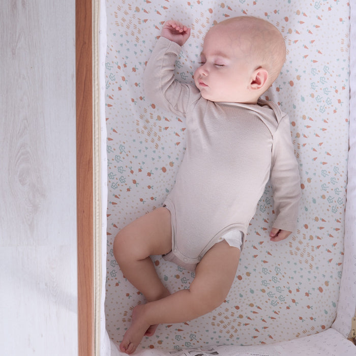 Overhead view of baby sleeping peacefully on the Good Life bedside crib fitted sheet in soft cream with vegetable garden print