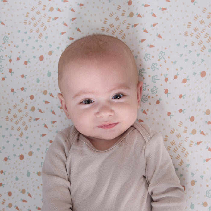 Close-up of baby smiling on the Good Life fitted sheet in cream with vegetable prints, blending warmth and whimsical character