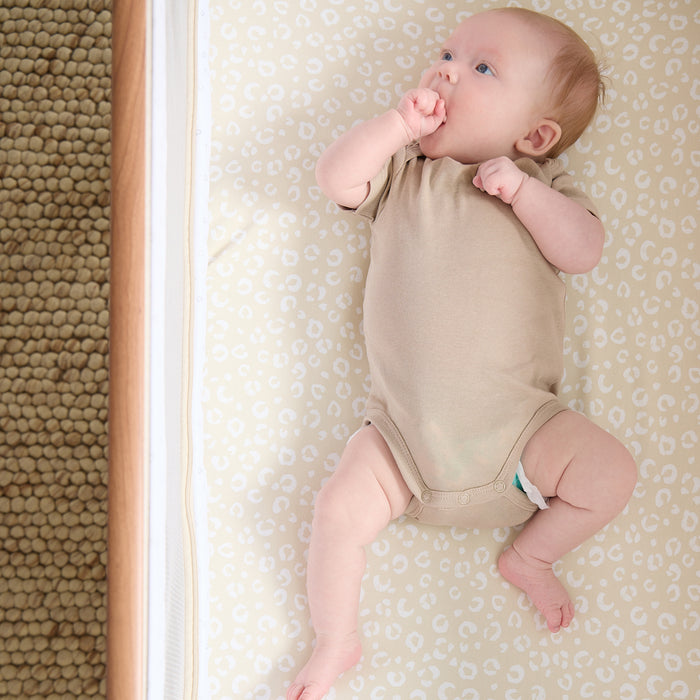 Overhead view of baby lying on the Love Cats fitted sheet in beige with delicate cat print, styled in a cosy bedside crib.