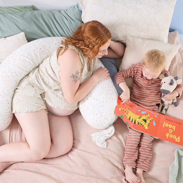 A woman, using the Good Life pregnancy pillow, and a child reading a book together on a bed