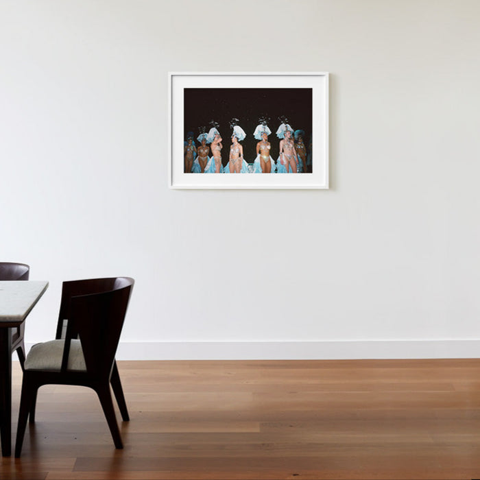Framed photograph of women in elaborate costumes hanging on a white wall above a wooden floor