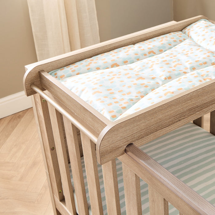 Close-up of the universal cot top changer in oak finish securely fitted to cot bed, featuring raised sides and a leopard print changing mat