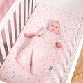 Baby lying in a cot bed wearing a sleep bag, on pink printed fitted sheets.