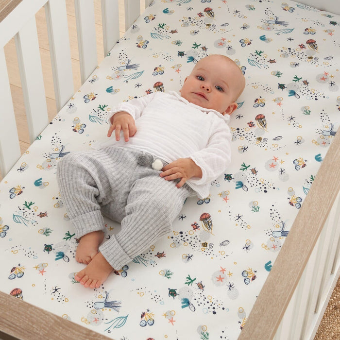 Baby lying on cot bed fitted sheet in Our Planet sea life print, highlighting soft organic cotton fabric for safe, gentle sleep