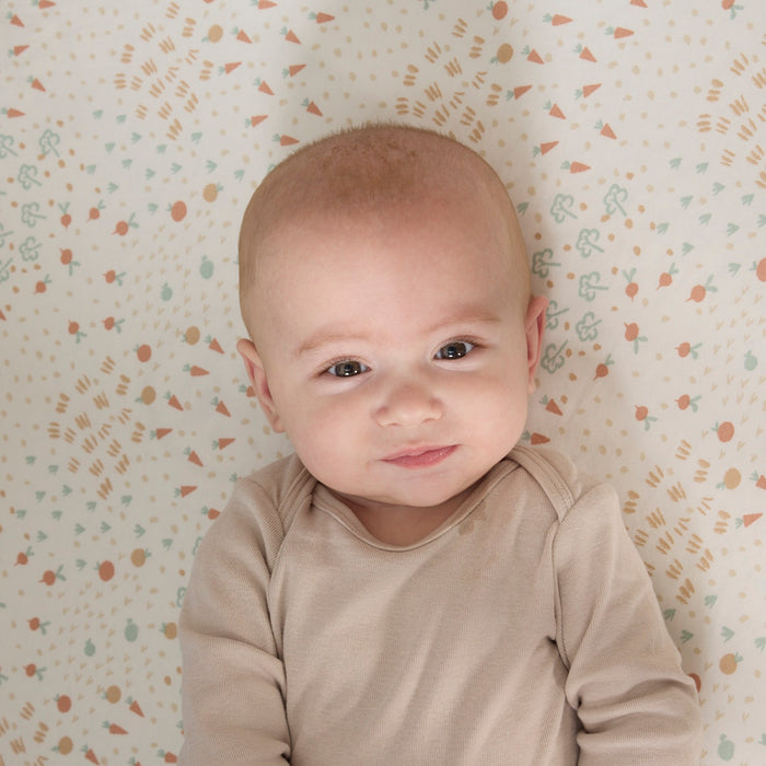 Baby wearing a beige onesie on a farm-patterned cot bed sheet from the Good Life collection.