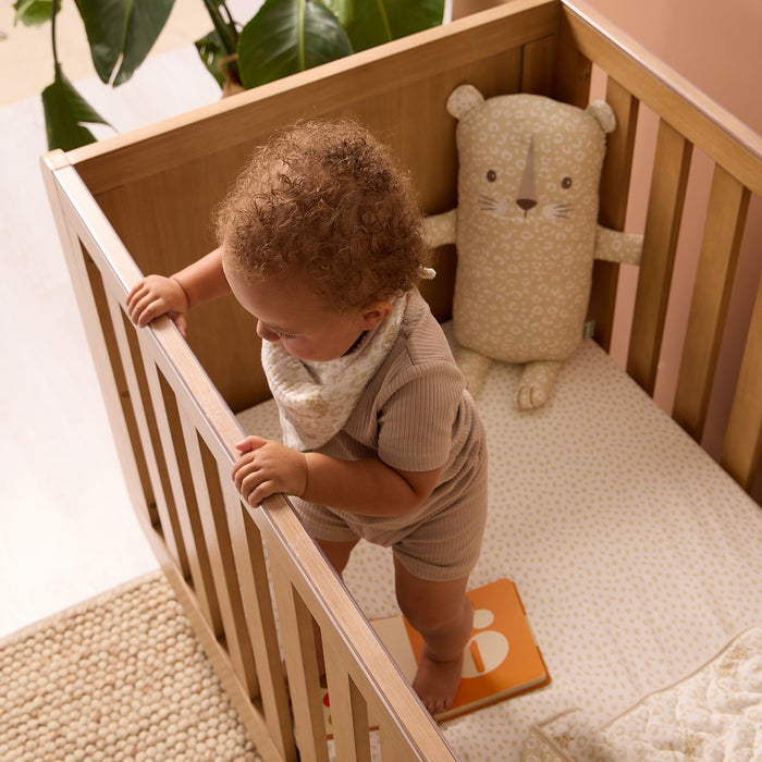 Overhead view of toddler standing in cot styled with Love Cats fitted sheet and toy accessories in warm beige tones and playful print.