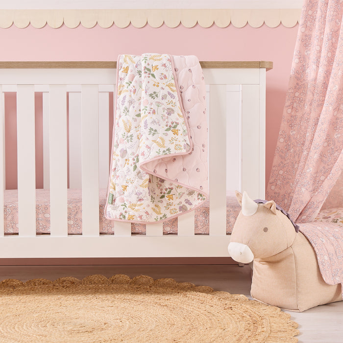 Tiny Tails cot bed coverlet draped over cot rail, featuring reversible print and strawberry pattern with pink piping