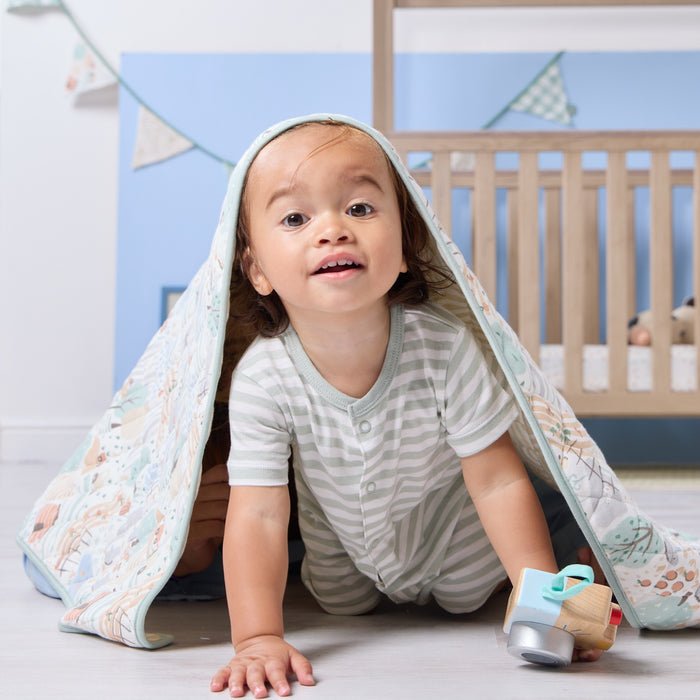 Toddler playing under the Good Life cot bed coverlet, smiling brightly while wrapped in its soft countryside inspired quilted fabric
