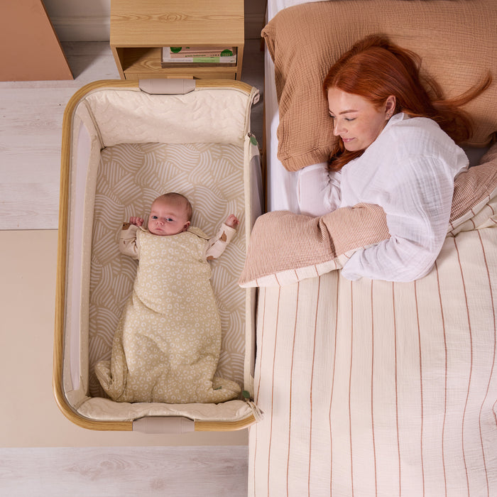 Overhead view of baby sleeping in the CoZee Air 2 bedside crib in toasted oak/sand positioned neatly beside the bed.