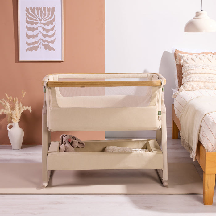 Front view of the CoZee Air 2 bedside crib in toasted oak/sand styled beside the bed, showing breathable mesh sides and storage shelf.