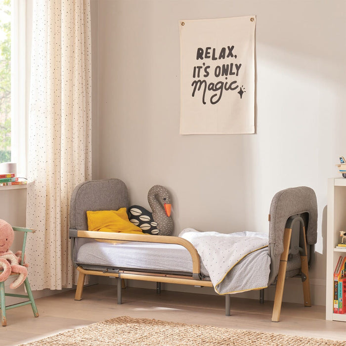 CoZee XL in oak and charcoal shown in toddler bed mode with padded ends, side guard, and soft grey upholstery in a bright nursery