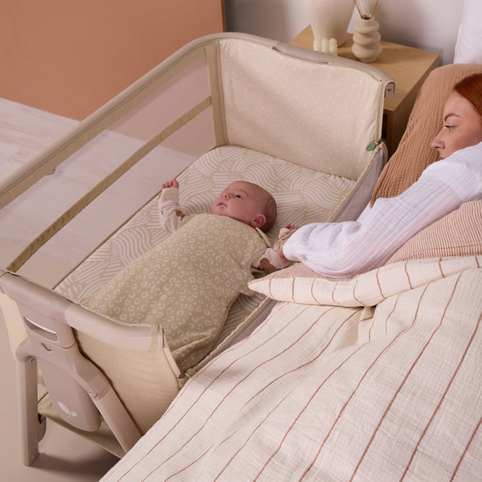 Baby resting beside parent in the CoZee Zen in sand bedside crib, highlighting safe co-sleeping positioning and breathable mesh sides.