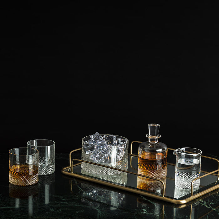 A gold tray holds Richard Brendon's Diamond Shot Glass Set of 2 with cut crystal design: a decanter with amber liquid, an ice-filled container, a water pitcher, and two empty glasses. Two more shot glasses—one with whiskey—rest beside the tray on a dark surface.