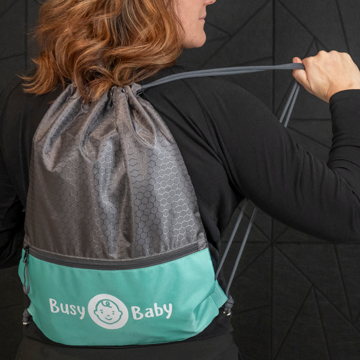 A person with shoulder-length hair wearing a black shirt is seen from behind, carrying a gray and turquoise Busy Baby Drawstring Backpack with the words "Busy Baby" and a baby face logo printed on it. Perfect for on-the-go parents, this Busy Baby gear stands out against the dark, textured panels in the background.