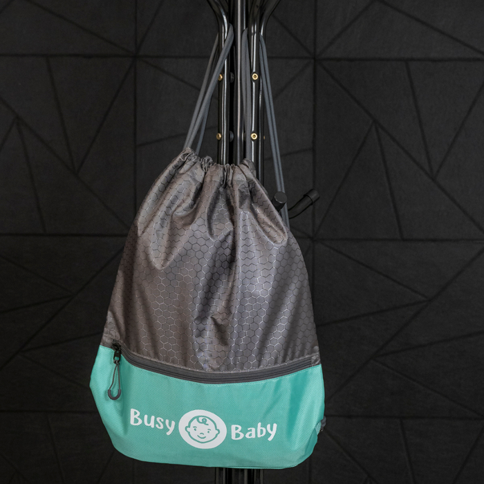 A high-quality Busy Baby Drawstring Backpack in gray and teal with a pattern of hexagons is hanging on a black metal rack. The bag features the Busy Baby logo and the text "Busy Baby" along with a smiling baby's face. The background consists of a geometric black wall.