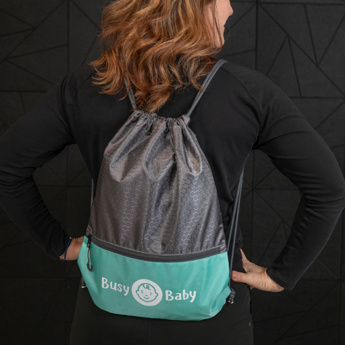 A person with wavy brown hair is shown from the back wearing a black long-sleeve shirt and a gray and turquoise Busy Baby Drawstring Backpack. The backpack, showcasing "Busy Baby" gear, features a baby face logo. The background is a dark, textured wall.