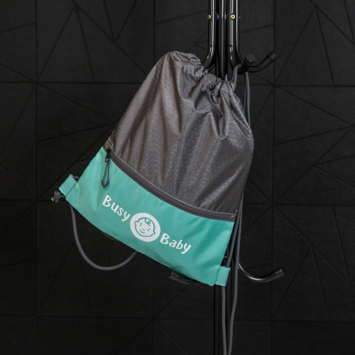 A high-quality gray and teal Busy Baby Drawstring Backpack with the text "Busy Baby" and a smiling baby face logo hangs on a black metal coat rack. The backdrop features a textured, geometric black wall, showcasing this essential piece of Busy Baby gear.
