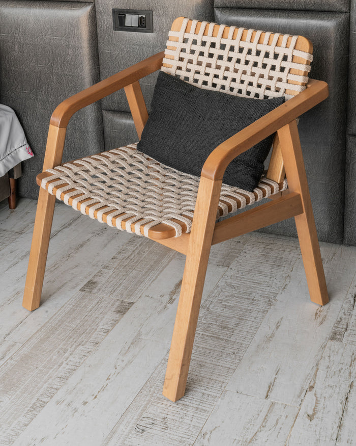 A wooden armchair with woven beige and white rope seat and back, styled with a dark gray throw pillow, set against a gray upholstered headboard and light wood floor.