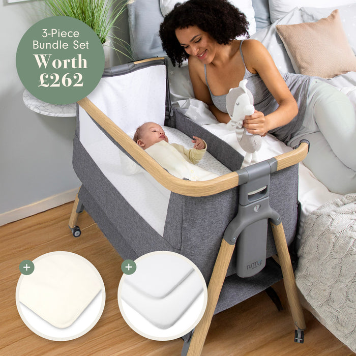 A woman with a baby in a grey crib with wooden legs, surrounded by baby items.