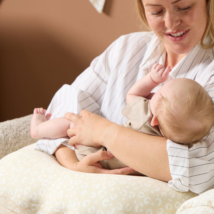 Mother cradling baby on the Love Cats feeding pillow in beige leopard print, demonstrating comfortable, supported feeding position.
