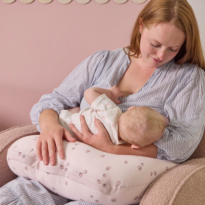 A woman breastfeeding a baby using the Tiny Tails nursing pillow in pink with a strawberry pattern, against a pink wall.