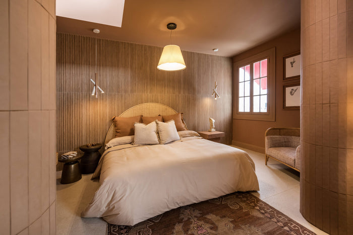 A modern bedroom featuring a bed dressed in cream bedding, set against a wall with vertical wood slats. The room has warm brown walls, a hanging conical pendant light, a patterned rug, and a small upholstered bench near a window.