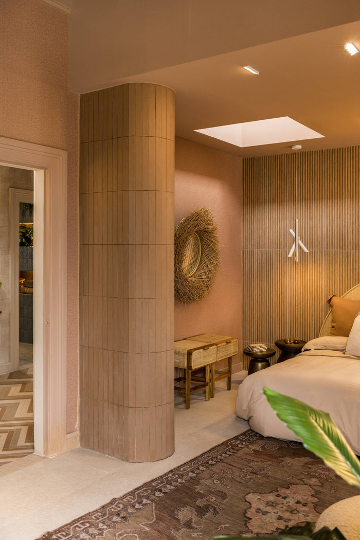 A modern bedroom interior with warm beige and blush tones, featuring a tall cylindrical column, a slatted wood accent wall, a bed with white bedding, and a patterned rug.