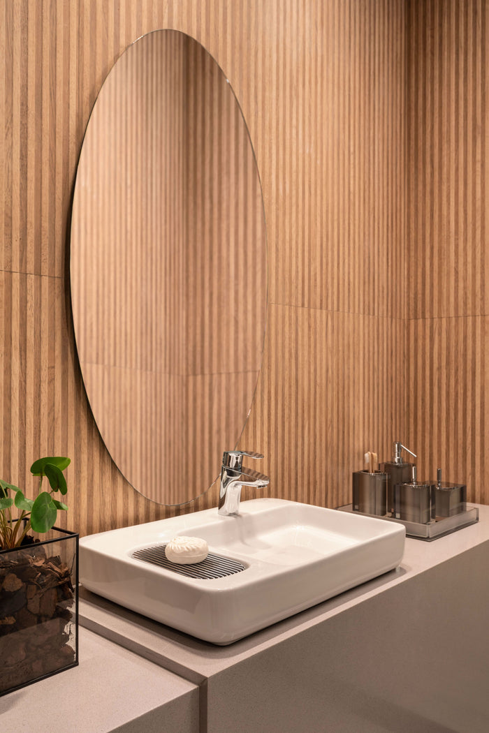 A modern bathroom vanity setup featuring a white rectangular vessel sink with a chrome faucet, a large oval wall mirror, and a light beige countertop against a wood-look paneled wall.