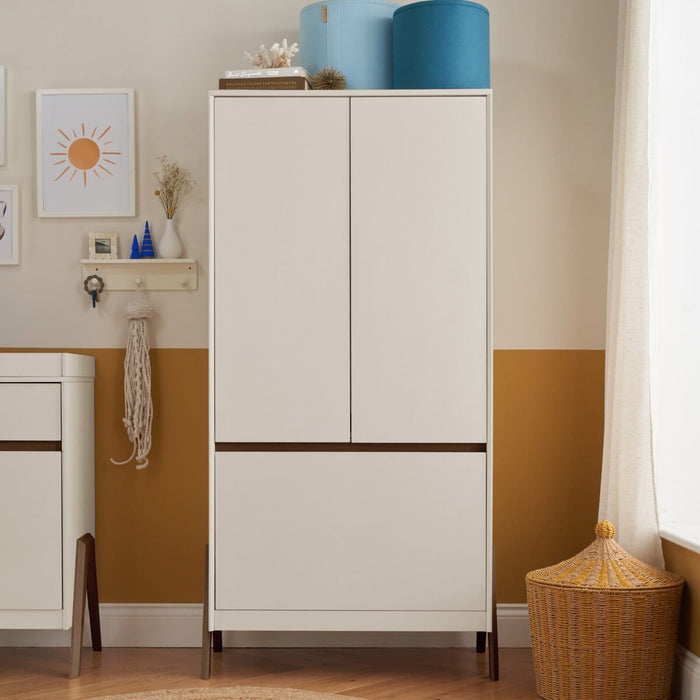 A modern white sand and warm walnut wardrobe with handleless doors and a hanging rail, placed in a room with a yellow wall and a wicker basket on the floor.