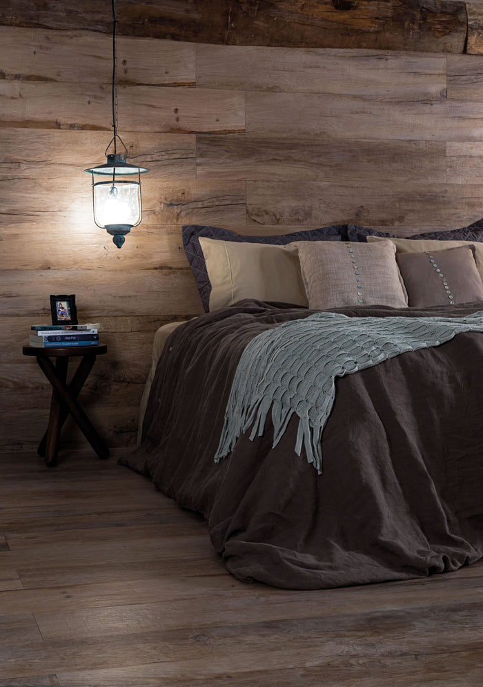 A rustic bedroom scene with a bed dressed in dark brown bedding and a light sage throw blanket, set against a wood-paneled wall with a hanging lantern light and a small side table.