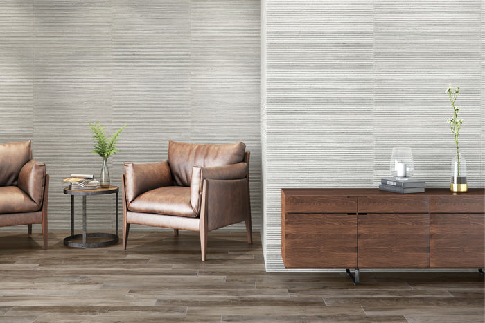 A modern interior scene showing textured light gray walls, medium-brown wood flooring, a brown leather armchair, a matching side table with a plant, and a dark wood credenza with decorative objects.