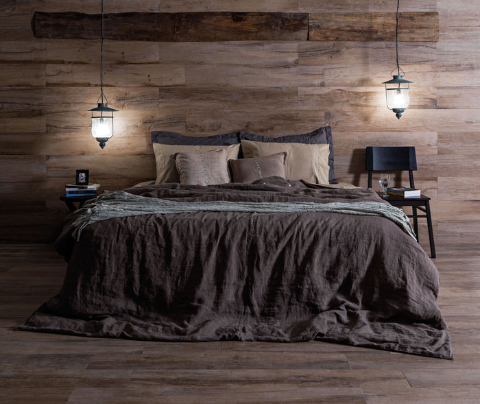 A rustic bedroom scene with a bed dressed in dark brown and beige bedding, set against a wood-paneled wall with two hanging lantern lights and a small side table on each side.