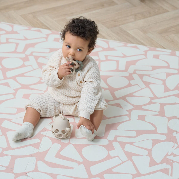 A baby sitting on a blush pink playmat with a geometric pattern.