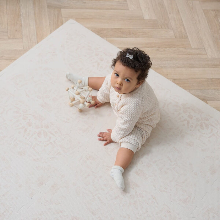 A baby sitting on a beige Infinity Baby Playmat, looking towards the camera with a toy in reach.
