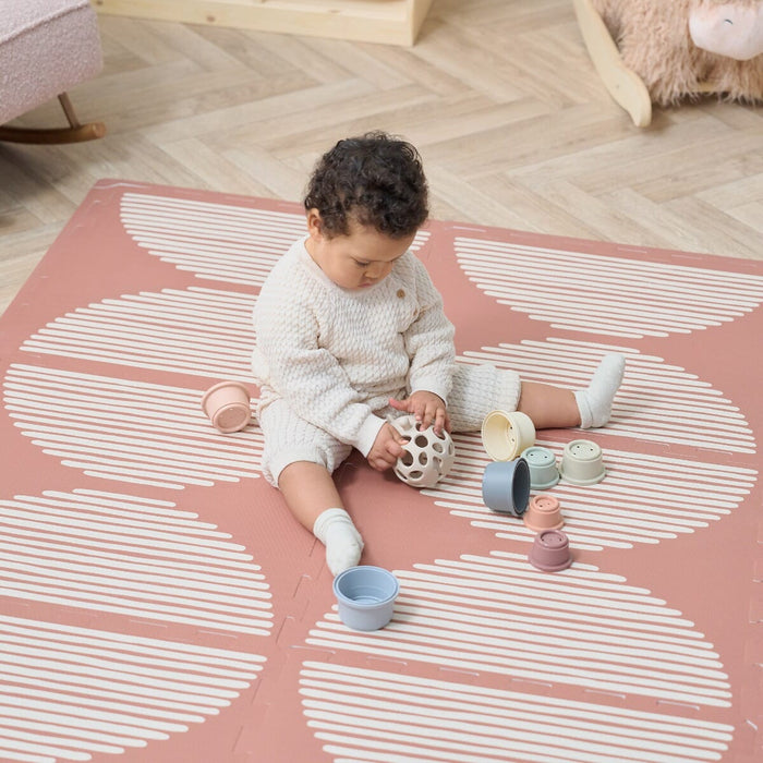 A baby sitting on an Infinity Baby Playmat with a geometric pattern in a warm nutmeg color, playing with toys.