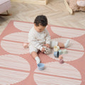 A baby sitting on an Infinity Baby Playmat with a geometric pattern in a warm nutmeg color, playing with toys.