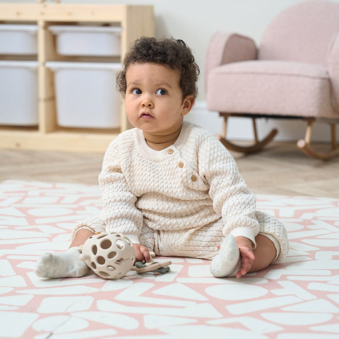 A baby sitting on the Oddity Blush Infinity Baby Playmat with a geometric pattern in a pink and white color, playing with toys.