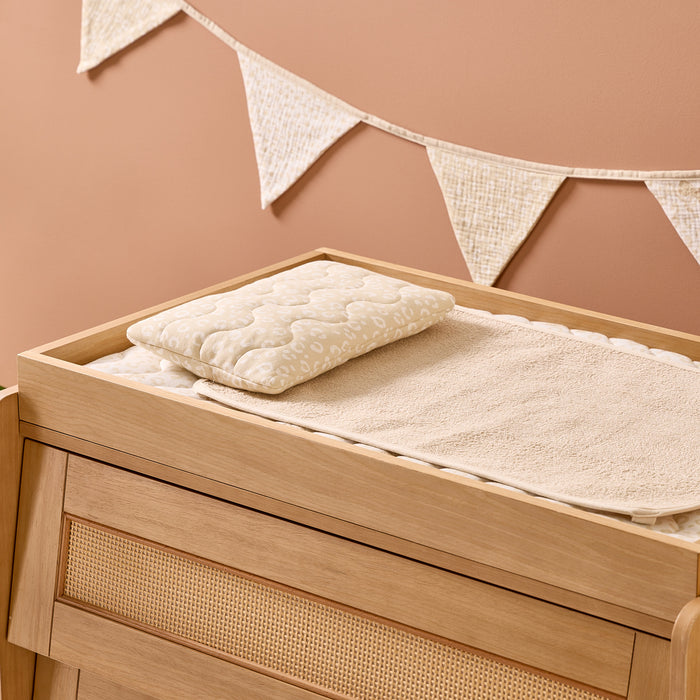 Wooden changing table with the Love Cats Luxury Changing Mat quilt and pillow against a beige wall with decorative flags.