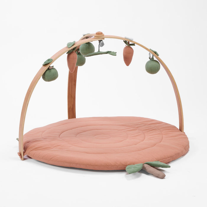 Circular muslin padded playmat in warm terracotta tone with wooden arch and hanging carrot and fruit toys.