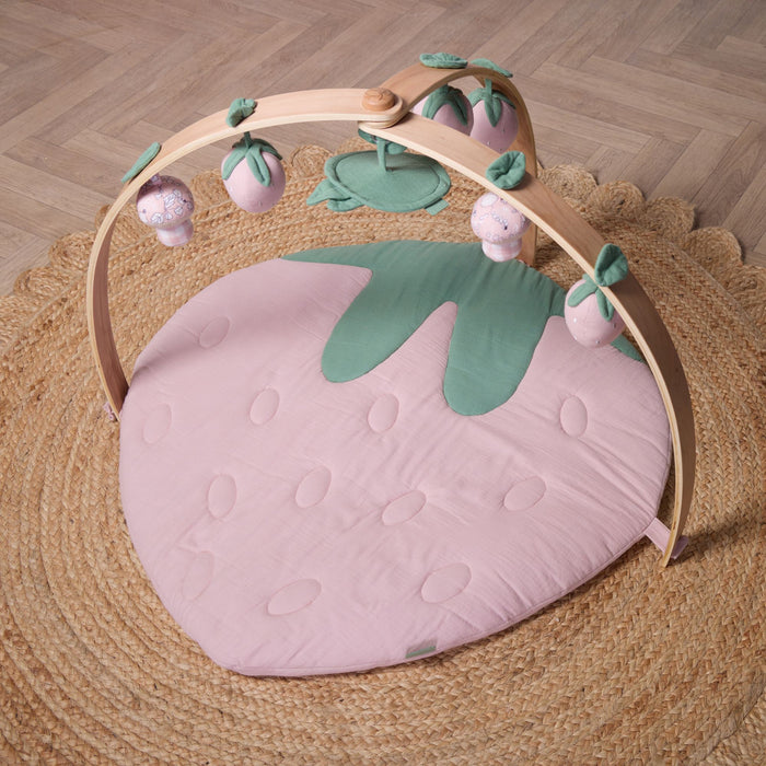 Strawberry muslin baby playmat with natural wood playgym arch and soft hanging toys, styled on woven rug.