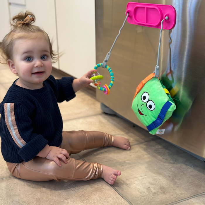 baby sitting on floor playing with toy attached to tether pink mini suction placemat on front of dishwasher