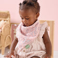 Baby wearing the Tiny Tails pink floral bib sitting on a chair with a pink background
