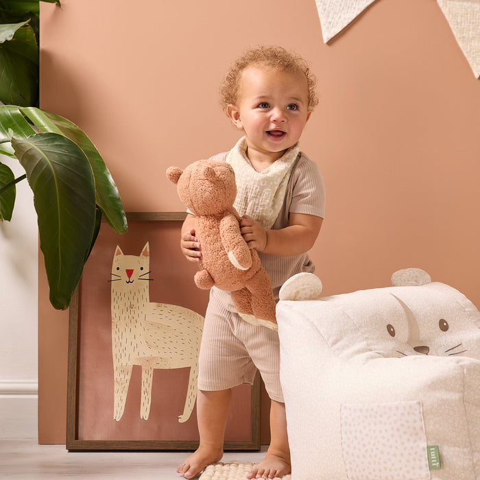 Child holding a teddy bear in a room with a wall featuring animal illustrations and a white chair.
