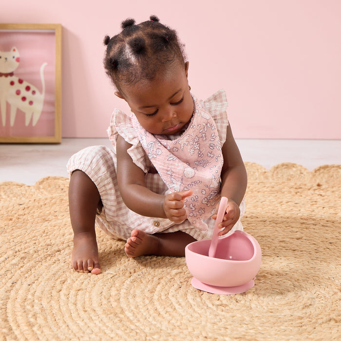 Baby wearing the Tiny Tails pink floral bib playing with a bowl on a room with a pink background