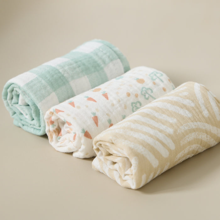 Three rolled Good Life muslin cloths in sage gingham, carrot print and neutral stripe, styled to highlight soft, breathable cotton texture.