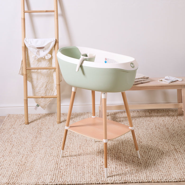 Nati Baby Bath in aloe on wooden stand with matching jug and storage shelf, styled in a calm neutral nursery setting.