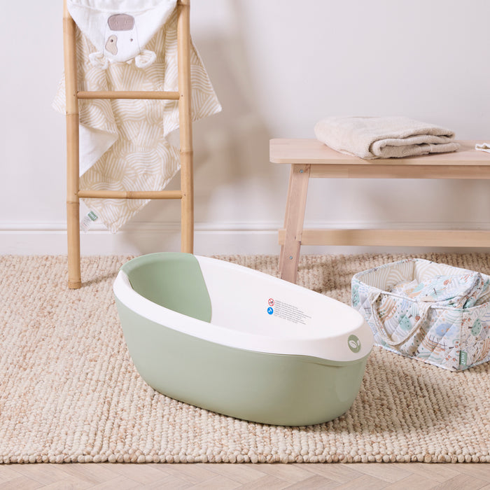 Nati Baby Bath in aloe styled on a woven rug with natural wood furniture and soft nursery accessories nearby.
